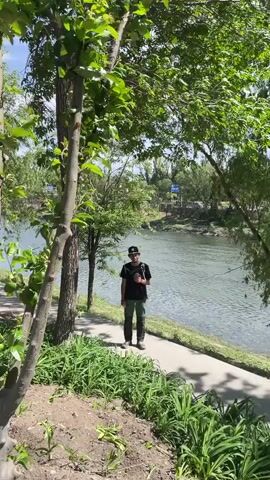 The video shows a man standing on a paved path beside a river. He is wearing a black t-shirt, green pants, and a baseball cap. He also appears to be carrying a bag or camera. The river is wide and flows from right to left. Trees line both sides of the path, providing shade. The day appears to be sunny.