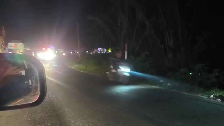 At night, a white van with red emergency lights passes by on a paved road. The van has a blue stripe and writing on the side. In the background are other vehicles, including an ambulance, also with emergency lights activated. The road is dark, reflecting the headlights of the vehicles. A side mirror of the car the video is taken from is visible. Bushes and trees line the side of the road.