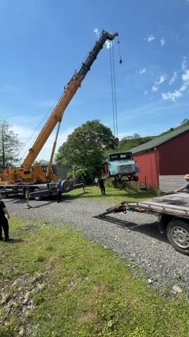 A crane is lifting a blue truck cab. A flatbed trailer is parked next to the cab. Several people stand nearby. The crane is yellow. A red barn is in the background. The setting is a yard with grass and gravel. The sky is blue with some clouds.