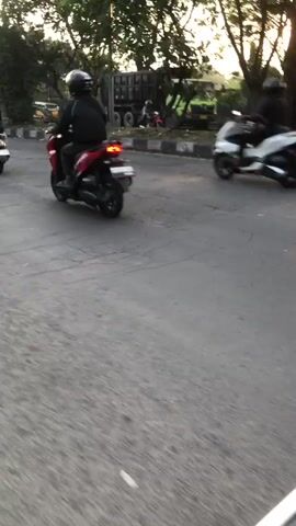 The media file captures a group of motorcyclists riding on a paved road in Ngaban, East Java. The scene appears to be set during the late afternoon or early evening, as indicated by the warm, golden sunlight filtering through the trees and casting long shadows. The sky is a soft gradient of light orange and grey, suggesting either sunrise or sunset.

The primary subjects are several motorcycles, each with a rider wearing protective gear, including helmets and jackets. The motorcycles are of various styles, with at least one being a scooter. The road is asphalt, showing signs of wear and tear with visible cracks and patches. Along the side of the road, there are trees with dense foliage and some vegetation, hinting at a semi-rural or suburban setting. In the background, structures that appear to be houses or buildings can be seen amongst the trees.

The activity taking place is a motorcycle ride, possibly a group outing or a journey. The motion blur of the motorcycles and riders indicates they are in motion, moving forward along the road. The overall mood seems dynamic and active, with a sense of camaraderie among the riders. There are no visible text elements in the media file. The weather appears clear and pleasant, conducive to outdoor activities like motorcycle riding.