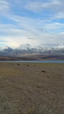 The video shows a wide shot of a dry, flat landscape in front of a large lake, with snow-covered mountains in the background. The sky is blue with scattered white clouds. The ground in the foreground is predominantly brown and grey, with small patches of sparse vegetation. In the distance, a body of water stretches across the horizon, reflecting a pale blue hue. Behind the lake, a range of mountains rises, their peaks covered in white snow. The lighting is natural, suggesting it's daytime. The camera is static.