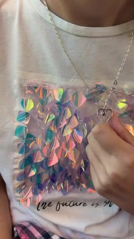 The video shows a close-up of a person wearing a white t-shirt with a colorful sequined design on the chest. A silver chain with a heart-shaped pendant rests on the t-shirt. A hand is shown reaching out and touching the sequined design. The person's lower body is partially visible, wearing a pink and white plaid garment. The background is not visible.