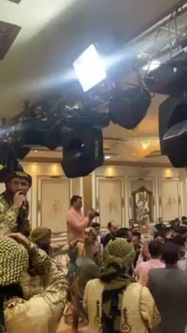 A festive atmosphere unfolds in what appears to be a banquet hall filled with a large crowd. The room is well-lit with overhead lighting fixtures and a chandelier, and the walls are decorated with ornate gold trim. People are gathered and moving, many clapping and dancing to the music, which includes singing into a microphone by someone wearing a traditional headdress. Several individuals in similar traditional attire are moving through the crowd. Someone is taking photos. Stage lighting is mounted on a truss system suspended from the ceiling.