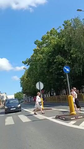 The video pans across a sunny street scene. A car is seen driving across a pedestrian crossing. On the left, a church with a red roof and a steeple is partially visible. People are seen walking along the sidewalk. In the foreground, there is construction fencing with yellow caution tape. Trees line the street. The sky is blue with some white clouds.