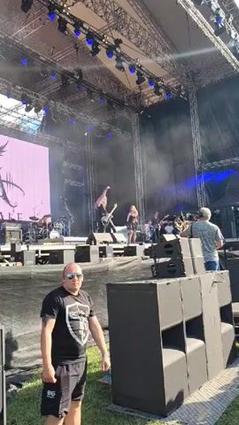 A man wearing sunglasses, a black t-shirt with a white logo, and black shorts stands on grass in front of a stage. Behind him, a band performs on the stage, with members playing instruments and singing. A large screen displays a graphic behind the band. Several large black speakers are positioned in front of the stage. The scene appears to be an outdoor concert or festival.