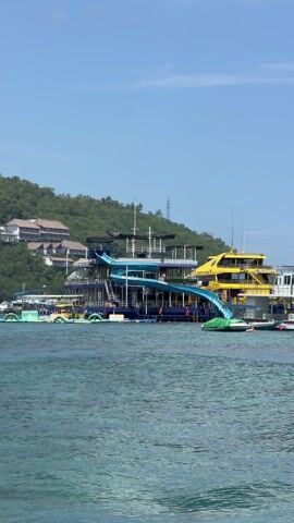 The video shows a stationary, multi-tiered, dark-blue boat with a large, curvy, light-blue water slide extending from its upper deck directly into the ocean, as well as an adjacent bright-yellow boat of similar height and design. The water in the foreground is clear, with slight ripples. In the background, a lush green hillside rises, dotted with several large houses. The sky is a clear, bright blue. The water slide boat has several inflatable floating toys connected to it.