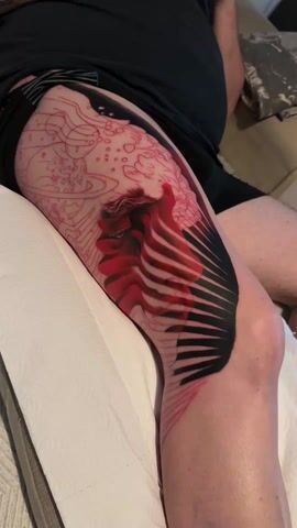 A person is getting a tattoo on their right thigh. The tattoo is a combination of red, black, and pink lines. The red and black parts of the tattoo appear to be more complete, while the pink lines are more like an outline of a design. The person is lying on a white surface. They are wearing a dark shirt and shorts. The tattoo features a side profile of a face with black lines radiating out from the face and red lines creating a striped pattern across it. A sketched, less-detailed design is near the top of the thigh.