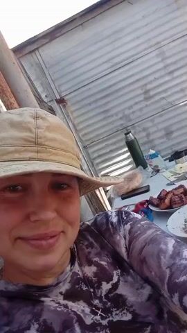 A woman wearing a tan sun hat and a long-sleeved gray and purple tie-dye shirt is sitting outside at a table. The table is covered with food, including what appears to be grilled meat, salad, cheese, and bread. There is also a thermos, a knife, a fork, and some snacks on the table. The woman is smiling and looking at the camera. The roof and part of the walls of the structure where the woman is are made of corrugated metal. A wooden wall is to the left. The lighting is bright and natural. The camera angle is low, giving a slightly upward view. The camera is held still for the duration of the short video.