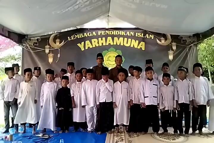 A group of around 18 young boys, likely students, stand on a stage under a white tent structure with a backdrop banner that reads "LEMBAGA PENDIDIKAN ISLAM YARHAMUNA". Two adult men stand in the center of the group. Most of the boys wear white shirts and black pants or robes, and the adults wear similar dark clothing and glasses. The group is posing for a picture, with some individuals raising their hands. The ground in front of the stage is covered with a blue plastic tarp, and the stage is set on a rug with a geometric design. The backdrop is decorated with cutout moons and lanterns. The background shows glimpses of trees and outdoor scenery.