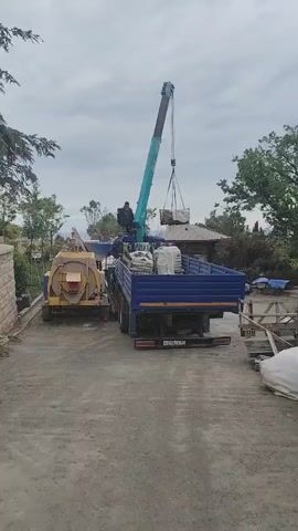 A blue truck, loaded with multiple sacks, is being unloaded by a blue and green crane, operated by a person sitting on top of the truck's cabin. The crane is lifting a large, wrapped rectangular object from the truck bed. Another person stands on the truck bed next to the sacks. The truck's license plate reads H258CK72. To the left of the truck is a yellow cement mixer. In the background, there are trees and what appears to be a small building or structure. The sky is overcast and gray. Debris, including pallets and more sacks, are to the right of the truck. The ground is dirt.