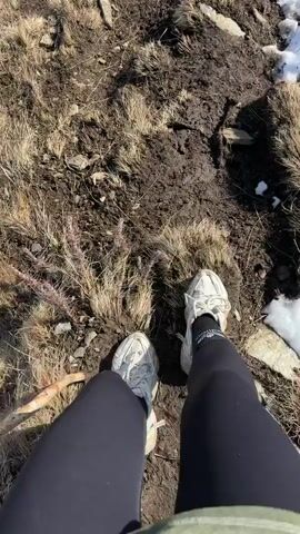 The video shows a first-person perspective of a person hiking on a mountain trail. The trail is muddy and covered in patches of dry grass, small rocks, and dwindling patches of snow. The hiker wears white sneakers and black leggings. As the view pans upward, a wide landscape is revealed, featuring a steep mountain slope dotted with trees and rocky terrain, leading down to a valley below with a glimpse of a city in the distance. The sky is visible at the horizon with a light blue hue.