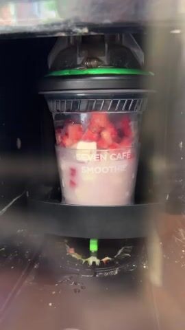 A clear plastic cup, labeled SEVEN CAFE SMOOTHIE, is situated inside a blending machine. The cup contains a light-colored base, likely yogurt or ice cream, with chunks of red fruit, possibly strawberries, and white pieces, possibly cream cheese or marshmallows, mixed in. The lid of the cup is secured and connected to the blending mechanism. The machine appears to be in operation, with a green nozzle visible below the cup, emitting a small amount of white liquid or residue. The surrounding area of the machine appears somewhat dirty and stained with green and yellow residue.
