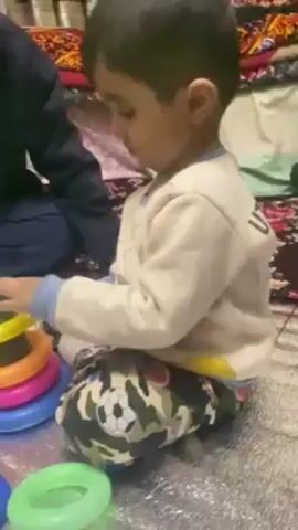 A young boy is sitting on the floor, playing with a ring stacker toy. He is wearing a white long-sleeved shirt with "UT AHA" printed on the back and camouflage pants with soccer ball designs. An adult is sitting beside him. The boy picks up a red ring and attempts to place it on the stacker, which is green and on top of a blue base. Various colored rings are scattered around him on a patterned rug.