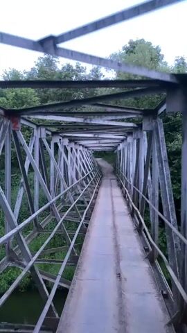 The video shows a first-person perspective walking across a metal truss bridge. The bridge is made of weathered gray metal, with a narrow pathway consisting of two parallel metal tracks. The pathway is flanked by metal railings. The bridge is surrounded by lush green trees and vegetation, suggesting a natural, possibly wooded environment. The sky above is a pale blue, transitioning to a darker blue in later frames, indicating it might be early morning or late evening. The camera moves forward, creating a sense of movement across the bridge. There are no other people or animals visible in the video. The overall impression is one of a serene, somewhat industrial structure situated within a natural landscape.