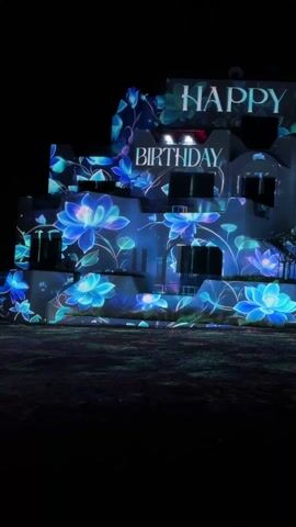 At night, an illumination display is projected onto the side of a building. The building is white, multi-storied, and features several windows. The projection includes the words "HAPPY" stacked over the word "BIRTHDAY," as well as images of stylized blue and turquoise flowers with green stems. A small tree is partially visible, providing some separation between the building and what appears to be a darkened yard in the foreground.  A black speaker on a stand sits to the right. The scene appears to be still and no people are readily visible.