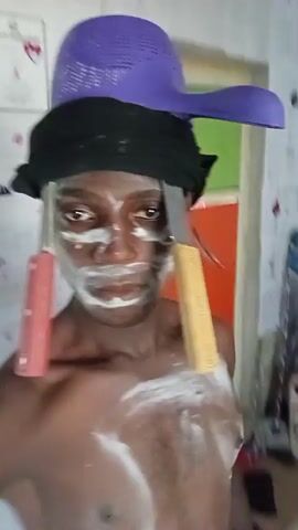 A topless person with brown skin, white facial makeup, and a purple colander on their head is holding the colander. They have a black cloth wrapped around their head and two knives stuck through the cloth on either side of their face. White paste is smeared on their chest. The background includes a door with a green panel, a red basket, and other items. The person looks directly at the camera.