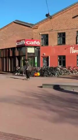 The video shows several young men skateboarding in an urban outdoor space on a sunny day. They are skating on a red-paved plaza with a low concrete ledge. One skater attempts a trick on the ledge. A brick building stands in the background, with signage indicating a café and entrance. Bicycles are parked near the building. Other people are seen sitting and standing around the plaza, some appearing to watch the skaters.