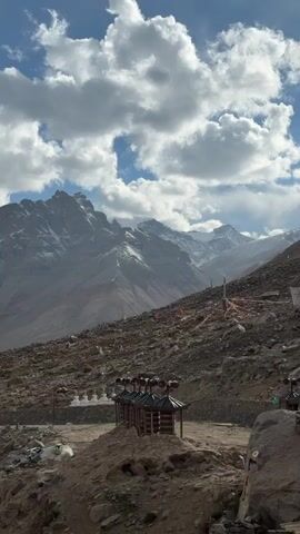 A slightly low-angle shot captures a mountainous landscape under a sky filled with white, puffy clouds against a blue backdrop. In the midground, a rocky, brown hillside slopes upwards. At the bottom of the hill, a series of small wooden structures are visible. Higher up the slope, prayer flags are scattered. Dominating the background are jagged, snow-capped mountains. A solar panel is placed in the foreground to the left. The overall scene is natural and rugged.