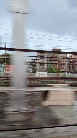 The video shows a blurry view from a train window. The train is moving, and the scenery outside is blurred. The video shows train tracks, overhead wires, trees, and buildings. There are also some signs visible, but they are too blurry to read. The video seems to be filmed during the daytime, as there is light outside. The weather appears to be overcast, with a cloudy sky. The train appears to be moving through a station or a town, as there are platforms and other trains visible. One train is green and white, and another is orange and red. There are also some freight cars visible, which are red and orange. The video is very short, lasting only about four seconds.