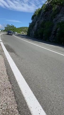 The video shows a daytime scene on a two-lane asphalt road curving through a mountainous or hilly landscape. The camera is positioned on the roadside, capturing vehicles as they drive past. The sky is bright blue with scattered white clouds, and the sun is shining. To the right of the road is a steep, rocky slope covered with some greenery and trees, while the left side shows a shoulder with gravel and a white solid line marking the edge of the road.

Several cars pass the camera:
1. A black Peugeot, possibly a 308, with license plate "SO 184-MP".
2. A two-tone grey and white Citroën C4 X, with a Turkish license plate "34 PSC 401".
3. A dark-colored sedan, which appears to be a Chrysler 300C.
4. A white Skoda, possibly an Octavia or Scala, with a partially visible license plate "SO 359-AL".
Additional cars can be seen in the distance, indicating ongoing traffic. The road appears well-maintained, with clear white lane markings. The overall impression is of a scenic drive on a clear day.