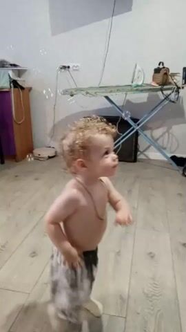 A young child with curly blonde hair and no shirt is standing on a wooden floor, looking up at a bubble machine that is being operated by an unseen person. The bubble machine is purple and green and is creating a stream of bubbles that are floating up towards a black television mounted on the wall. The child is wearing patterned shorts and white socks. In the background, there is an ironing board with an iron on it, and various household items. The room appears to be a living room or playroom, with neutral-colored walls and wooden flooring. The overall scene is one of play and amusement.