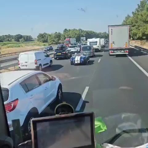 The video captures a roadside view from inside a vehicle, with a focus on a classic blue and white convertible sports car driving on a highway amidst traffic. The sports car has white racing stripes and a driver wearing a helmet. Other vehicles visible include a black SUV, a white caravan, a semi-truck, and several other cars and vans. The environment appears to be a sunny day with trees and vegetation visible in the background. The footage is shaky, suggesting it was recorded while moving.