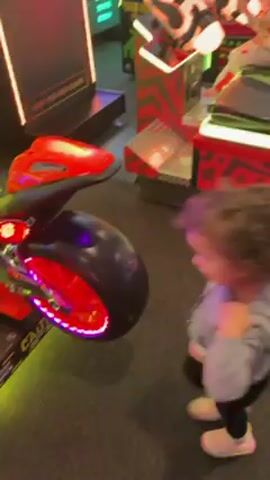 A toddler-aged child with curly hair and wearing a gray hooded sweatshirt, black leggings, and white sneakers is excitedly playing next to a red and black motorcycle arcade game in a brightly lit arcade. The child is initially standing next to the game, gesturing with their arms and looking at the screen. The camera moves to capture the arcade game with its bright neon lights and visible game screen. The child then walks around the game before being picked up by an adult whose arm and torso are visible. The arcade is filled with other games and people in the background.