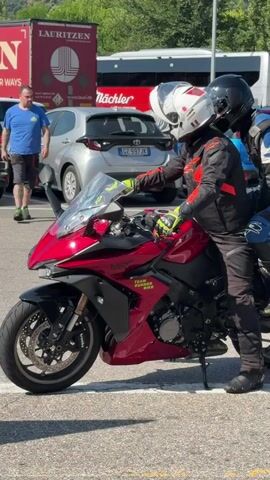 In Villa, Italy, on a bright, sunny day, two individuals are positioned on a red and black sportbike. The rider in front wears a white helmet, black and red motorcycle jacket, and black pants, with yellow gloves. They are operating the motorcycle, which has a prominent windshield and "TEAM RUNNER BIKE" written on its side. Behind the rider, a passenger sits on the motorcycle, wearing a black helmet and a blue and black motorcycle jacket.

The scene appears to be a parking lot, indicated by the paved ground and parked vehicles in the background. A silver car with a license plate starting with "CZ" is visible directly behind the motorcycle. Further in the background, a large red truck with "LAURITZEN" and "WE WAYS" in white lettering is parked, along with a bus that has "Nöchler" written on it. The overall setting suggests an outdoor location, possibly a rest stop or a gathering point for motorcyclists. The sunlight suggests it is daytime.