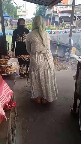 A woman in a light green headscarf and a long, patterned dress stands with her back to the camera, facing a food stall. She is looking towards another woman who is wearing a black headscarf and a black dress with gold embellishments. The woman in black is standing behind the food stall, which displays skewers of meat. There is smoke rising from a grill behind the woman in the patterned dress, suggesting the food is being cooked. The scene appears to be outdoors, possibly on a street or market, with various structures and vegetation visible in the background.
