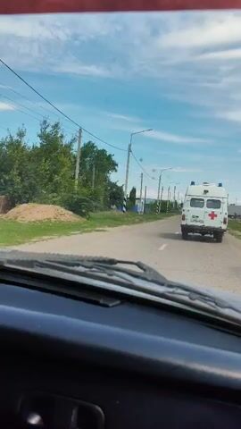 The video shows an ambulance with a red cross on its side driving down a road. It appears to be a rural area with houses and trees visible. The sky is blue with some clouds. The video is filmed from inside another vehicle, looking out through the windshield. The camera also captures a reflection of a person's hand holding a phone in a side mirror.