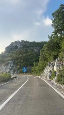 The video shows a paved road winding through a mountainous, forested area. The road has white lane markings and a guardrail on the left side. A blue sign with white text and a map is visible on the right side of the road. The sky is partly cloudy with the sun shining. The overall environment appears to be rural and scenic.