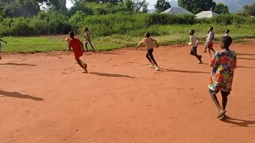 The video shows a group of about ten children playing soccer on a reddish-brown dirt field. The children are wearing a variety of clothes, including T-shirts, shorts, long-sleeved shirts, and even a dress. Some are barefoot, while others are wearing shoes. A soccer ball is in play. The field is bordered by green grass and dense bushes and trees. In the background, a few houses can be seen. The sky is bright and sunny, suggesting it is daytime. The camera pans slightly to follow the movement of the children as they play.