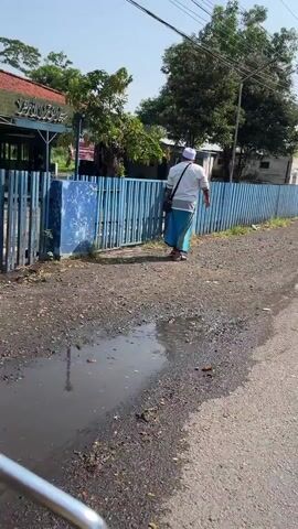 The video shows a man walking away from the camera on a dirt and asphalt road. The man is wearing a white shirt, a white cap, and a light blue sarong. He is carrying a dark bag over his right shoulder. He is walking alongside a blue metal fence. To the left of the fence, there is a building with a red roof and signs on it. To the right of the fence, there are trees and more buildings in the background. The ground in front of the fence has puddles of water and debris. The sky is clear and the sun is shining.