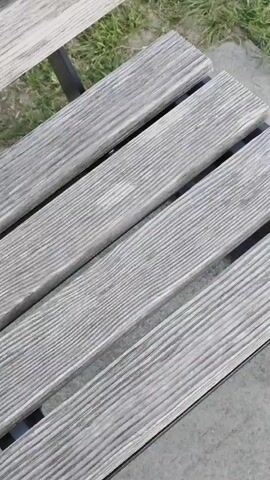 The video opens with a close-up of a weathered wooden park bench with a metal frame, suggesting it is outdoors. The bench is situated on what appears to be a paved area next to a grassy field. As the camera pans upwards and to the right, the field becomes the primary focus, showing a well-maintained expanse of green grass. Further in the distance, a dense line of dark green trees forms a treeline against a backdrop of rolling hills or mountains. The sky above is overcast with a muted gray hue, indicating potentially cloudy or late afternoon conditions. There are no people or animals visible in the footage.