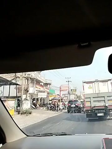 The video shows a street scene with several vehicles. In the foreground, a truck with a container is visible from the rear. To its left, a black SUV is stopped. Further to the left, on the side of the road, there are several motorcycles parked and some people are standing around them. Buildings line the street, with shops and signs, including an "Alfamart" sign. The road appears to be paved, and there are power lines and utility poles visible. The weather appears to be clear and sunny.