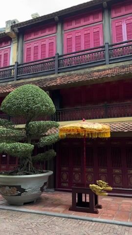 The video shows the courtyard of a building, possibly a temple or palace. The building has two levels, with red walls and doors adorned with gold details. There are balconies on both levels with black railings. A bonsai tree in a white pot sits on the left. Two ornate golden parasols stand near a bench with a canopy decorated with gold fringe. The courtyard is paved with red bricks. There are no people visible. The music playing suggests a peaceful and possibly spiritual setting. The camera pans slowly to the right.
