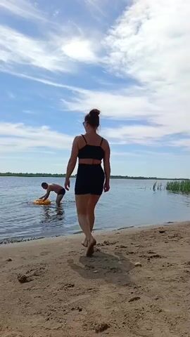 A woman in a black bikini walks towards the water on a sandy beach. She is facing away from the camera. In the water, a man is kneeling and holding a yellow inflatable ring. The scene is a body of water, likely a lake or river, with a distant shoreline visible. The sky is blue with scattered white clouds, suggesting it is daytime and fair weather. The overall mood appears relaxed and summery, indicative of leisure activities. There is no visible text in the media.