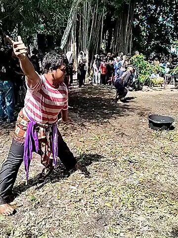 The video shows two men engaged in a whip fight in a rural area, possibly as part of a cultural or traditional performance. A crowd of onlookers surrounds the arena, suggesting that this event is of significance to the local community. The arena itself is a dusty, open space with two large metal bowls placed in the center. One man wears a striped shirt and a purple sash, wielding his whip in a wide, forceful arc. The other combatant is dressed in darker clothing and is actively trying to defend himself from the blows. The background is filled with trees and various plants, indicative of a tropical or subtropical climate. The scene is dynamic with the men moving and their whips flying, creating a sense of both danger and spectacle.