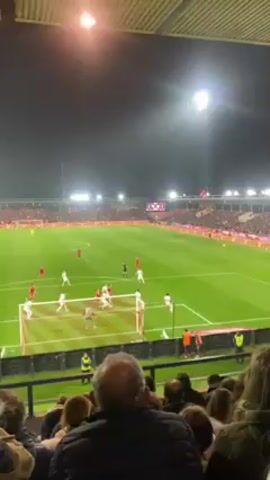 The video shows a soccer game from the perspective of a spectator in the stands. The game is being played at night under bright stadium lights. The field is green and well-maintained. Players in red and white uniforms are visible on the field. The ball is in play near one of the goals. The stands are filled with fans watching the game. The crowd is making noise, and a cheer can be heard as the ball goes into the goal. The video is shot from a slightly elevated position, giving a good view of the entire field. The quality of the video is decent, and the key actions are clearly visible.
