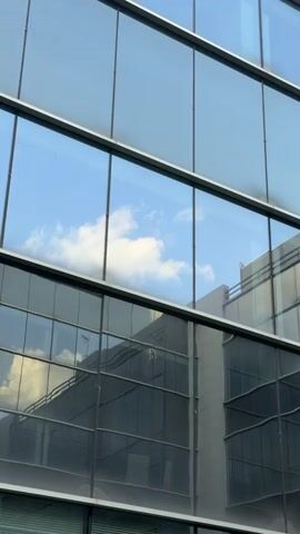 The media file displays the exterior of a modern, multi-story building primarily composed of large, rectangular glass windows. The windows feature dark, thin frames that create a grid-like pattern across the facade.

The glass is highly reflective, capturing elements of the surrounding environment. In the upper sections, a clear blue sky with scattered white cumulus clouds is prominently reflected, creating a serene visual effect. The lower sections of the windows reflect parts of an adjacent or opposite building, which appears to be a light-colored structure with its own dark, rectangular window openings and some visible rooftop or upper-level architectural features, including what looks like a balcony or railing.

The scene is brightly lit, indicating daytime. No people or specific activities are visible. The setting is an urban environment, specifically San Sebastián De Los Reyes, Spain. There is no discernible text within the image.