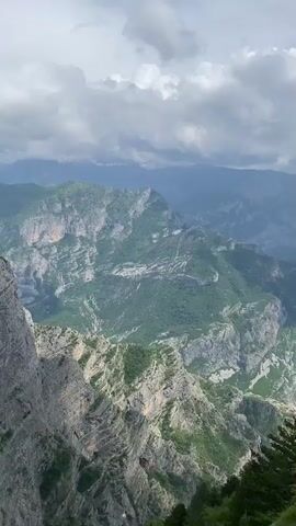 The video presents a panoramic view of a vast, rugged mountain landscape in Korita, Montenegro. The camera slowly pans from left to right, revealing layers of green and grey mountains under a cloudy sky. In the foreground, a steep drop-off features rocky slopes and scattered green trees and bushes. As the camera pans, a winding road can be seen snaking through a valley in the mid-ground, indicating human presence in this remote terrain. Distant peaks are visible, partially obscured by mist or clouds. Some small clusters of buildings or settlements are faintly visible in the valleys as the view shifts. The overall impression is one of majestic natural beauty and immense scale. The sounds of distant conversation can be heard, suggesting people are present at this viewpoint, admiring the scenery. The time of day appears to be daytime, with natural light illuminating the landscape despite the overcast conditions.