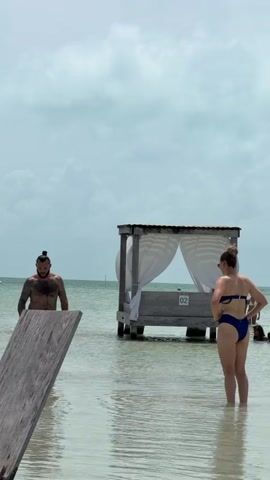 The scene shows a shallow body of water under an overcast sky. In the midground, a man with a beard and hair tied back is partially submerged on the left, facing right. To his right, a woman in a blue bikini stands facing away from the camera, with her hands on her hips. Between them and slightly behind, a wooden structure resembling a canopy bed or cabana, with white curtains and the number "02" visible, stands in the water. Further in the background, other people are partially visible in the water. A wooden structure is partially visible in the bottom left foreground.