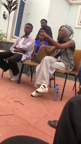 A group of people are sitting on chairs in an outdoor setting. The area is paved with red bricks. The people are dressed in casual clothing. A woman wearing a headscarf and patterned pants is talking and gesturing with her hand. A young man in a striped shirt is sitting next to her with his arms crossed. A water bottle is on the ground next to each of them. Behind them, two other individuals are standing. A plant in a pot is next to the building. A bicycle is visible behind the seated individuals. The building has a light blue facade.