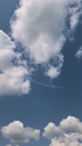 An airplane performs an aerial maneuver, leaving a smoke trail in the clear blue sky filled with fluffy white clouds.