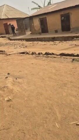 The video shows a dirt road in a rural area, with modest houses lining the street. The houses appear to be constructed of concrete blocks or a similar material and have corrugated metal roofs. There are some drainage ditches along the side of the road, made of concrete blocks. A person dressed in dark clothing and carrying what seems to be a rifle walks down the street in the middle of the video. The area looks dusty and somewhat impoverished, with trash scattered on the ground. There are also power lines visible overhead.