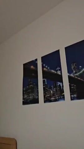 The camera begins aimed at the ceiling of a room, revealing a light fixture. It then pans down to show a wall with a two-part picture of a bridge and city skyline. The camera pans around the room, showing a desk with a gaming console, a black chair, and a partially opened doorway that reveals a bathroom and hallway. There are several pairs of slippers lying on the wooden floor in the doorway. An infant makes cooing noises throughout the video.