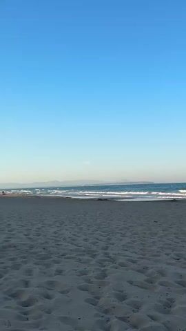 The video shows a sandy beach with calm ocean waves reaching the shore under a clear, bright blue sky. In the distance, there is a faint outline of hills or mountains on the horizon. The beach is made up of a light gray or tan sand, marked with numerous footprints. The ocean water is a turquoise color, transitioning to a deeper blue as it extends further from the shore. The waves are small and gentle. The sky is largely cloudless, providing a vast and open backdrop to the scene. There's a faint hint of a color gradient in the sky near the horizon. No people are visible in the shot.
