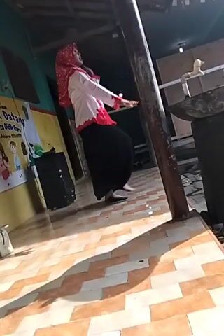 A person wearing a red hijab, white shirt with red trim, and a black skirt is dancing. The person is in a room with a tiled floor and a wooden pillar. There is a poster on the wall and a speaker in the corner. The person is moving their arms and body to the rhythm of the music.