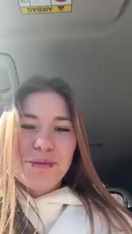 The video features three young women in the back seat of a car. The woman in the foreground, wearing a white hoodie and a seatbelt, is holding the camera. Her long brown hair is parted and hangs down her chest. Behind her, another woman is visible holding up her phone as if taking a selfie or recording something. She wears a black and white jacket. The third woman sits further back and is partly obscured. Outside the car window, there is scenery consisting of trees and a hint of sunlight. The car's interior includes the ceiling and headrests of the front seats. The women seem to be on a road trip or outing.