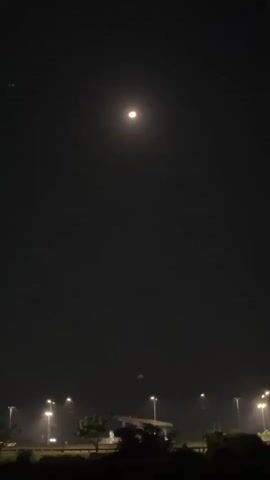 The media file is a vertical video recorded at night in Wunut, Indonesia. The scene depicts a dark sky illuminated by a bright, reddish moon. Below the moon, a faint, blurry green light is visible. In the background, a line of streetlights stretches across the frame, suggesting a road or an open area. Silhouetted trees and a structure, possibly a building or a bridge, are visible beneath the streetlights, indicating a semi-urban or a well-lit rural setting. The overall atmosphere is quiet and serene, with the moon being the central and most striking subject. There is no visible text in the video. The time of day is clearly night, and the weather appears to be clear enough to see the moon prominently, though there might be some atmospheric haze contributing to the moon's reddish hue and the blurry appearance of the green light.